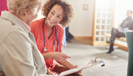 Doctor holding clipboard talks to patient, seated in waiting area.